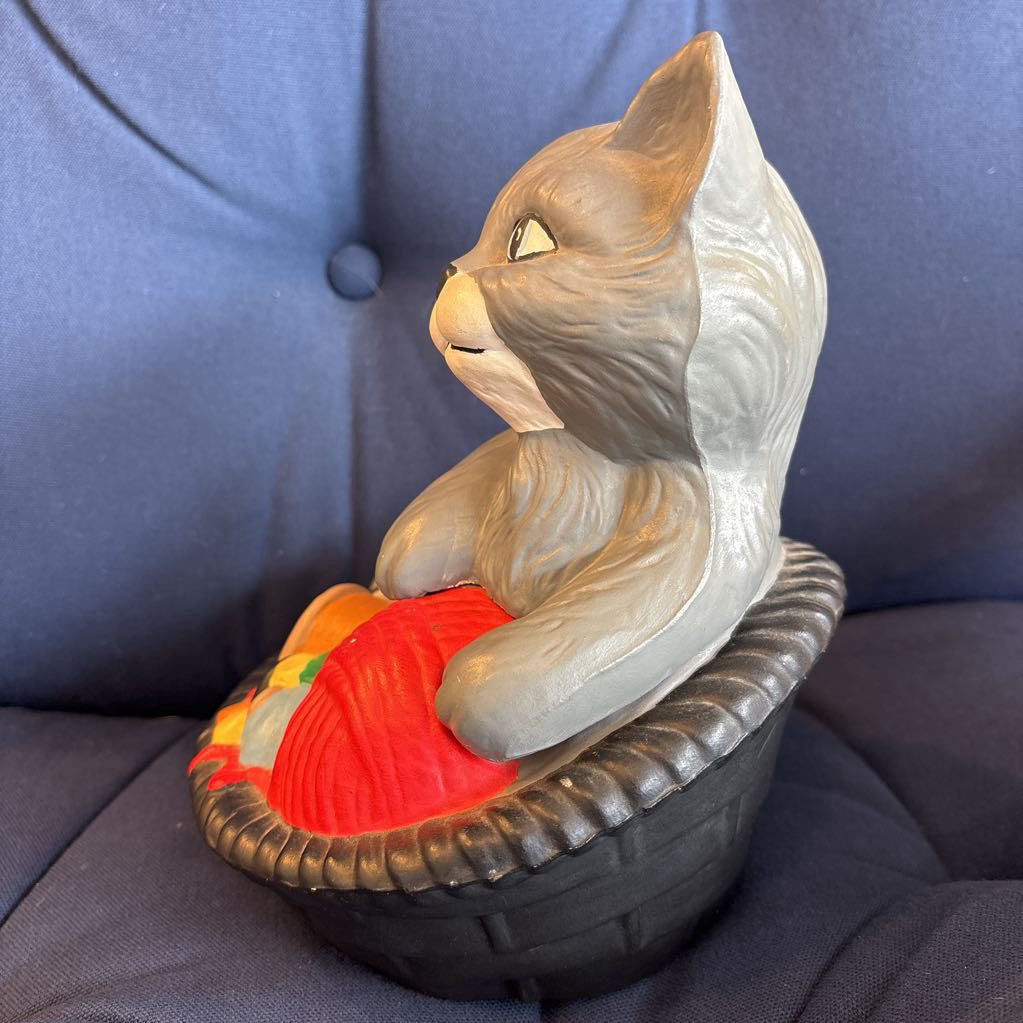 A ceramic figure of a gray cat in a bowl filled with yarn and sewing supplies sitting on a plush navy cushion. 