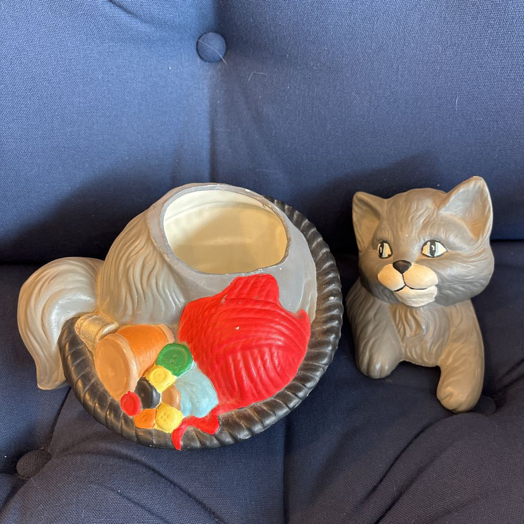 A ceramic figure of a gray cat in a bowl filled with yarn and sewing supplies sitting on a plush navy cushion. 