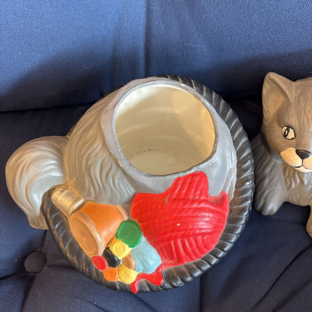 A ceramic figure of a gray cat in a bowl filled with yarn and sewing supplies sitting on a plush navy cushion. 