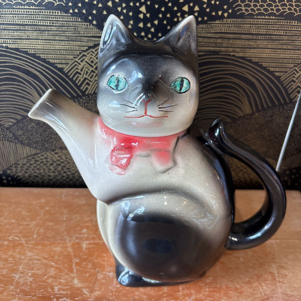 A ceramic teapot of a tuxedo cat with green eyes and a pink bowtie. Its arm is the spout and its tail the handle. It sits on a wooden surface against a black and gold wall. 