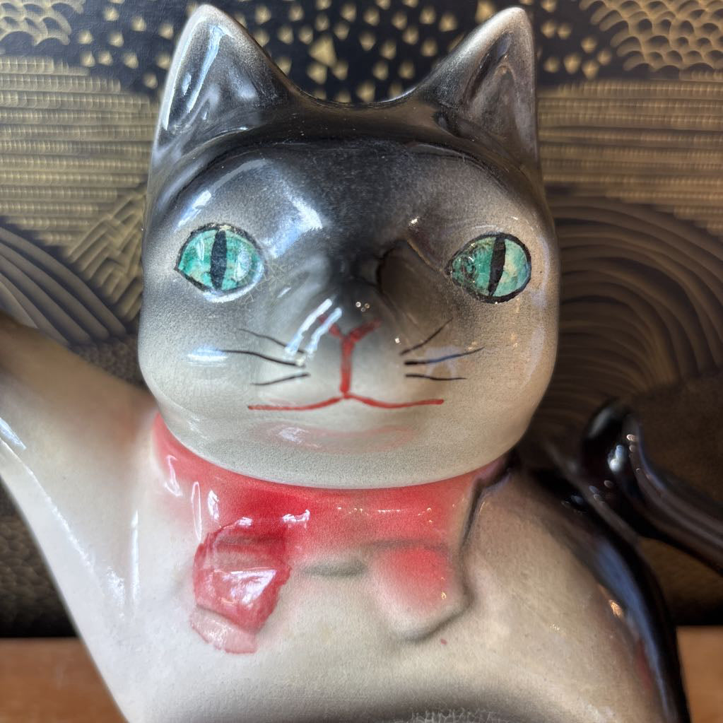 A ceramic teapot of a tuxedo cat with green eyes and a pink bowtie. Its arm is the spout and its tail the handle. It sits on a wooden surface against a black and gold wall. 