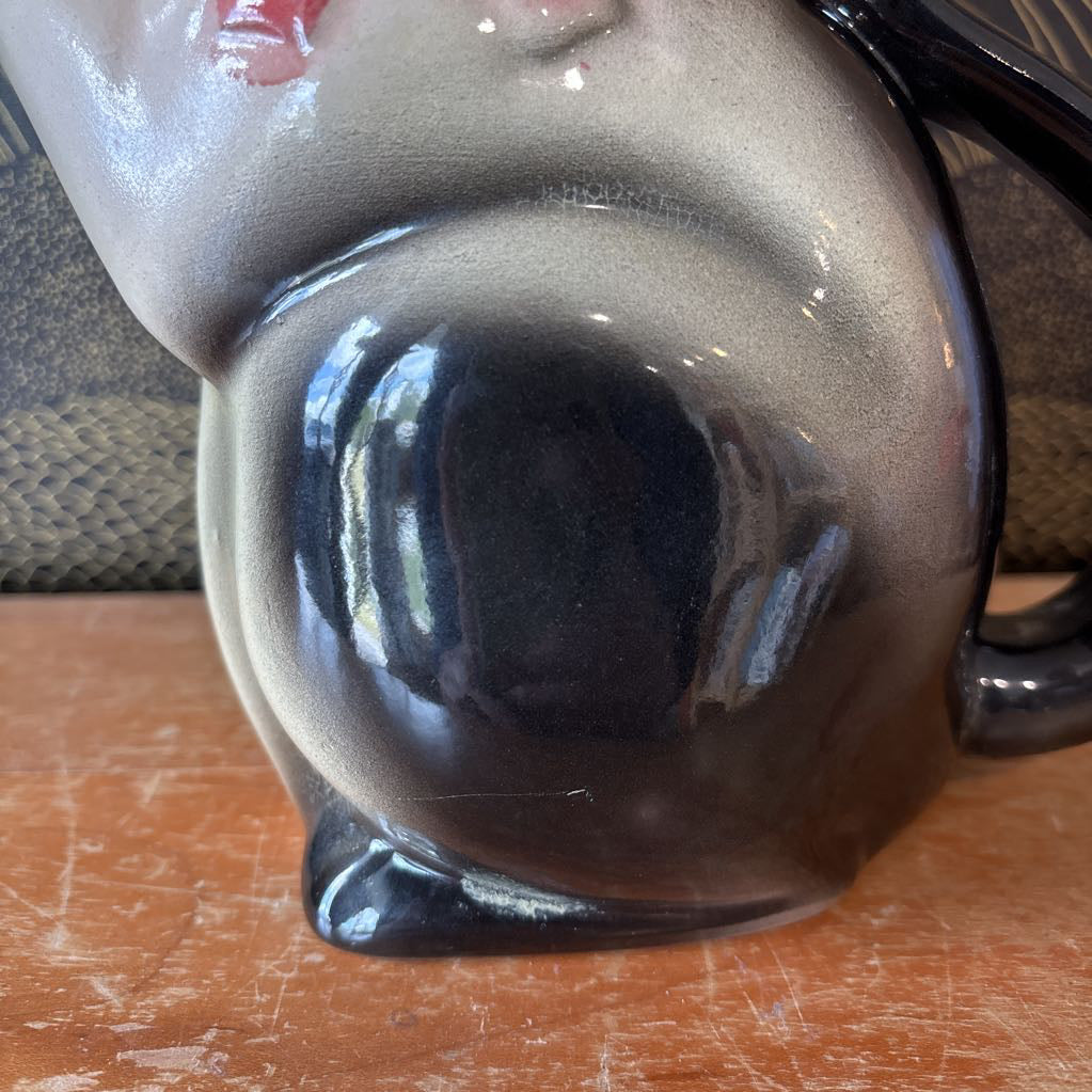 A ceramic teapot of a tuxedo cat with green eyes and a pink bowtie. Its arm is the spout and its tail the handle. It sits on a wooden surface against a black and gold wall. 