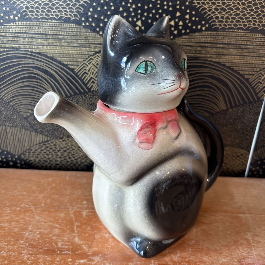A ceramic teapot of a tuxedo cat with green eyes and a pink bowtie. Its arm is the spout and its tail the handle. It sits on a wooden surface against a black and gold wall. 