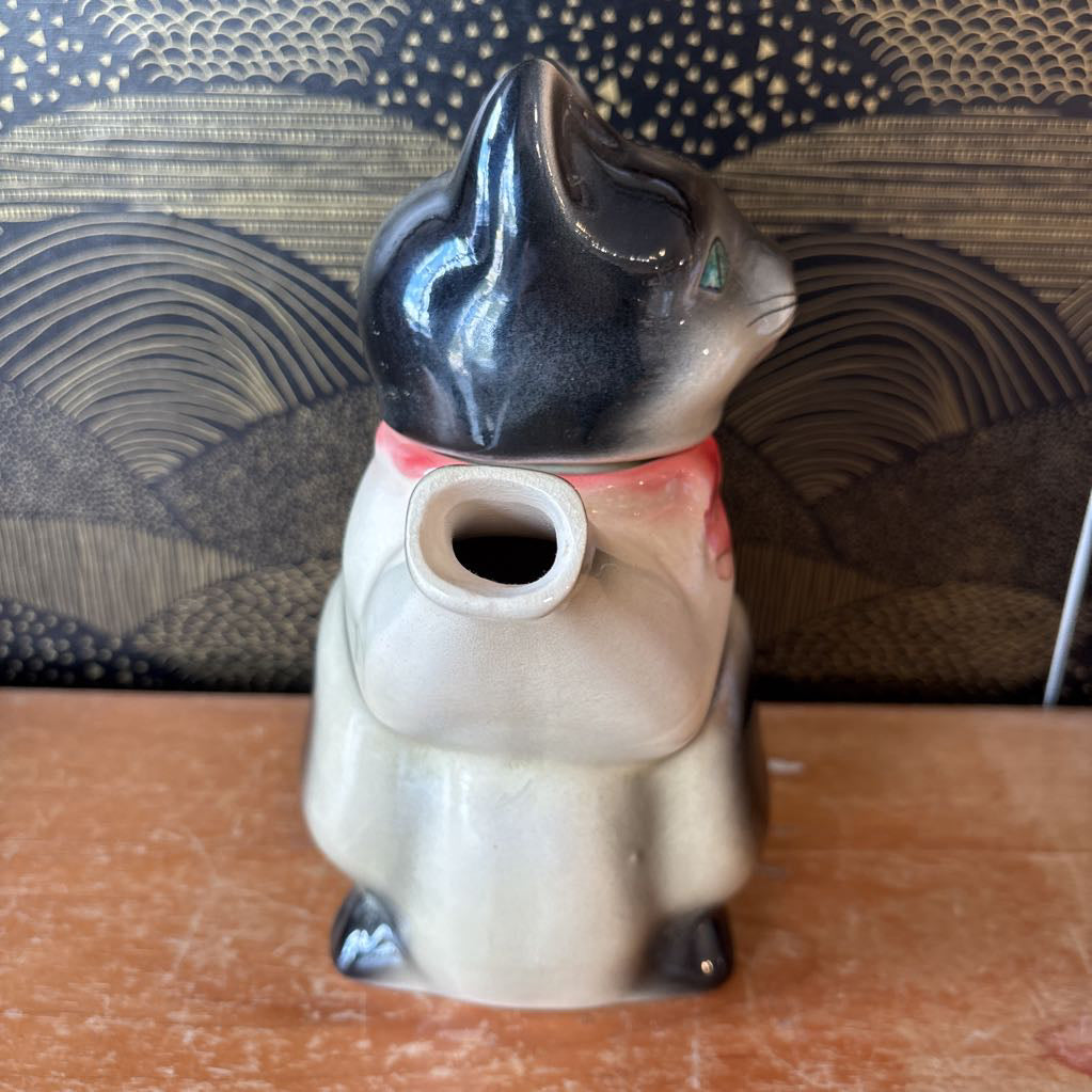 A ceramic teapot of a tuxedo cat with green eyes and a pink bowtie. Its arm is the spout and its tail the handle. It sits on a wooden surface against a black and gold wall. 