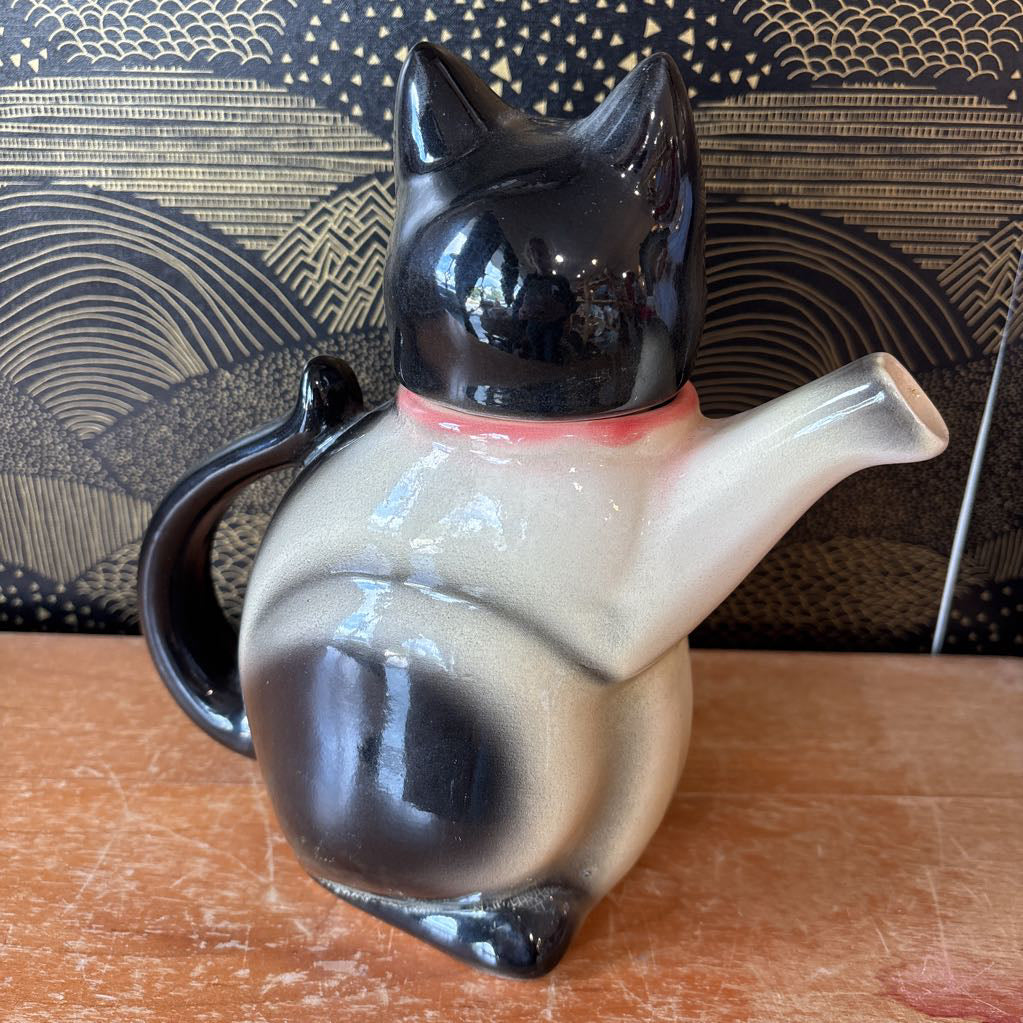 A glossy ceramic cat-shaped teapot or pitcher with a black head and tail, a cream body, and a small pink collar, shown on a wooden surface in front of a patterned black and gold background.