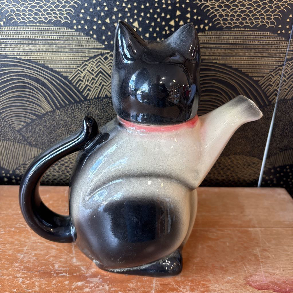 A glossy ceramic cat-shaped teapot or pitcher with a black-and-white gradient body, black handle and head, and a small pink collar, sitting on a wooden surface in front of a patterned backdrop.