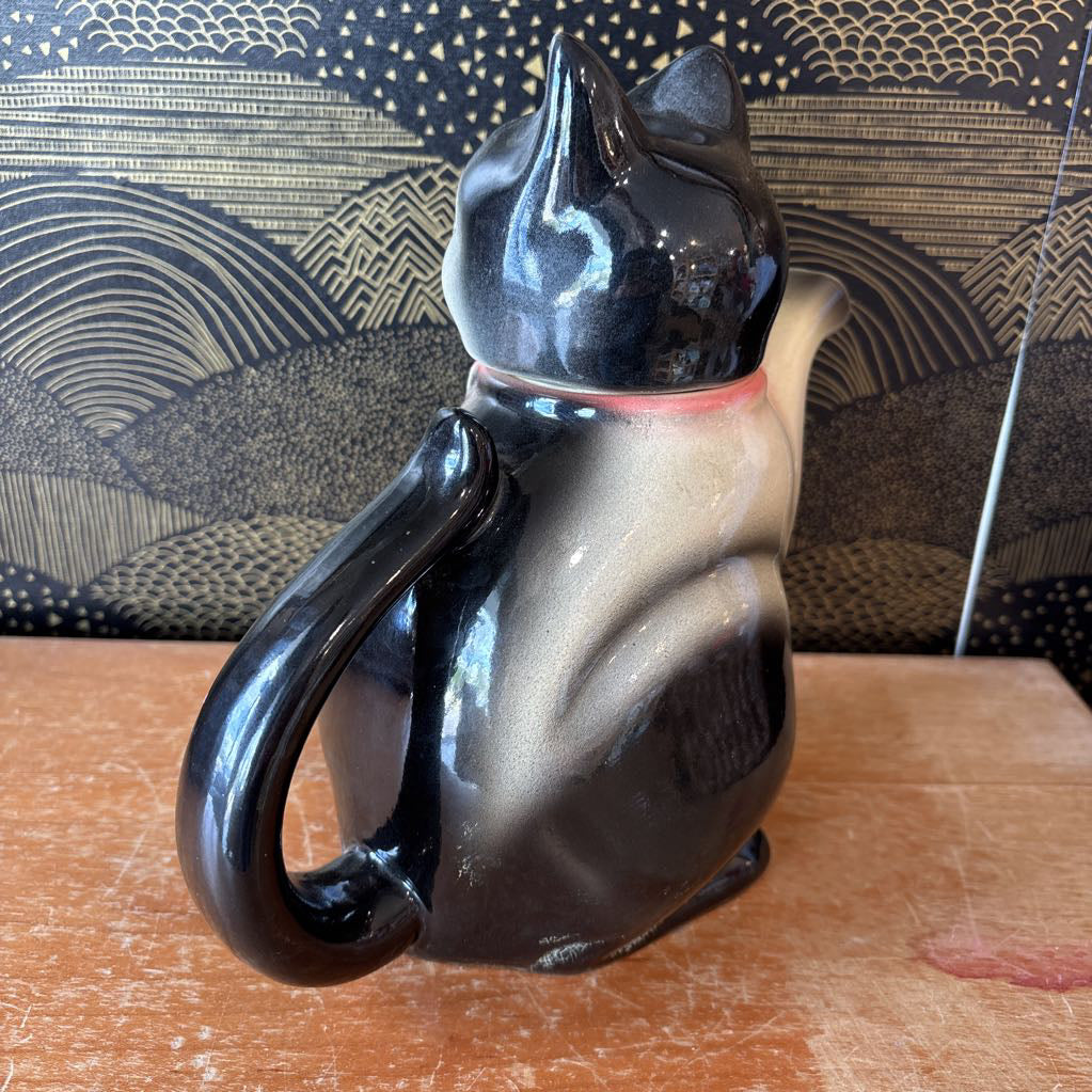 A ceramic teapot of a tuxedo cat with green eyes and a pink bowtie. Its arm is the spout and its tail the handle. It sits on a wooden surface against a black and gold wall. 