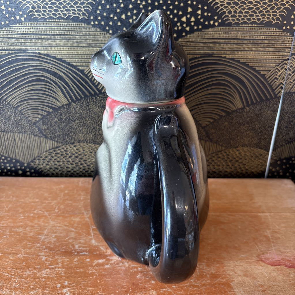 A glossy black cat-shaped ceramic pitcher with a red collar detail, sitting on a wooden tabletop in front of a patterned black and gold background.