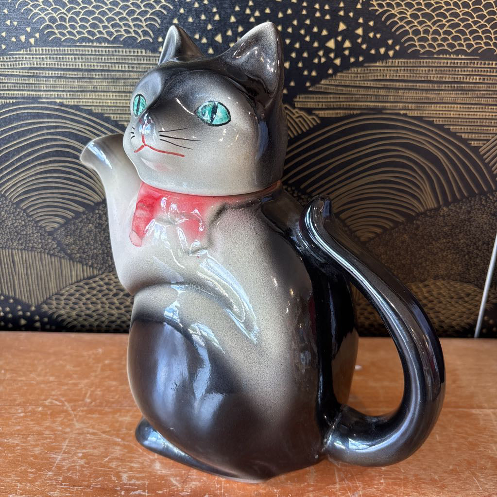 A glossy ceramic cat-shaped pitcher or teapot with a gray and black glaze, turquoise eyes, and a pink collar, shown on a wooden surface against a patterned dark background.