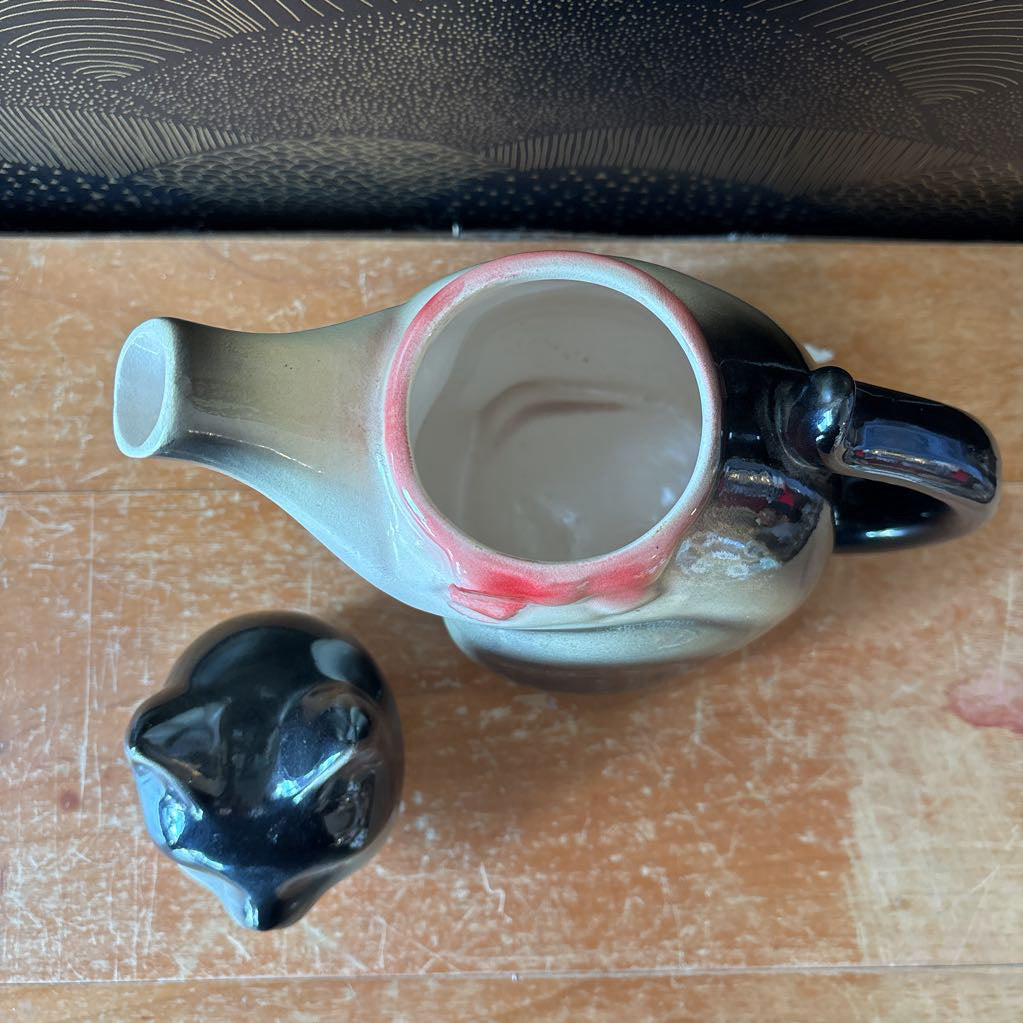 A ceramic cat-shaped teapot with its lid placed beside it on a wooden tabletop. The teapot has a glossy black handle and lid, with a body that transitions from black at the top to a warm beige/cream at the bottom, accented by a pink interior rim.