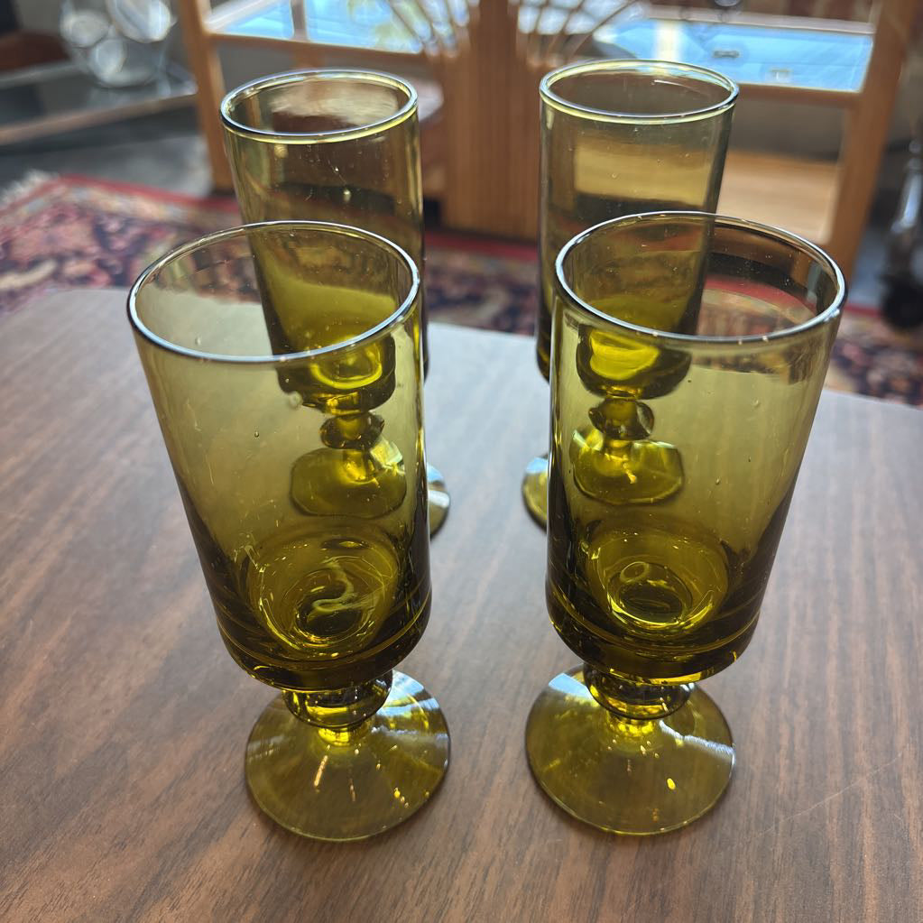 Four green glass tumblers with footed bases arranged on a wooden table in an indoor setting.