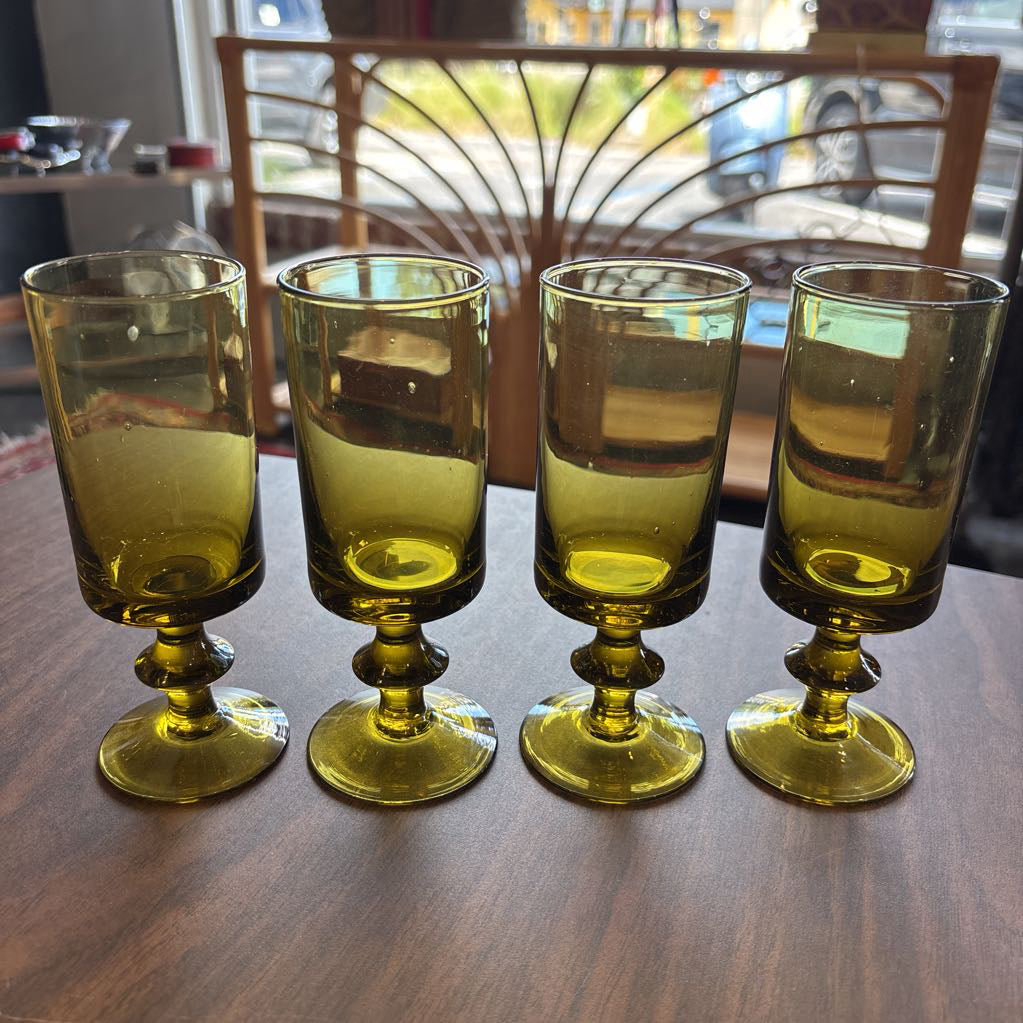 Four vintage olive green glass goblets with pedestal bases arranged in a row on a dark wooden table, with a blurred indoor background.