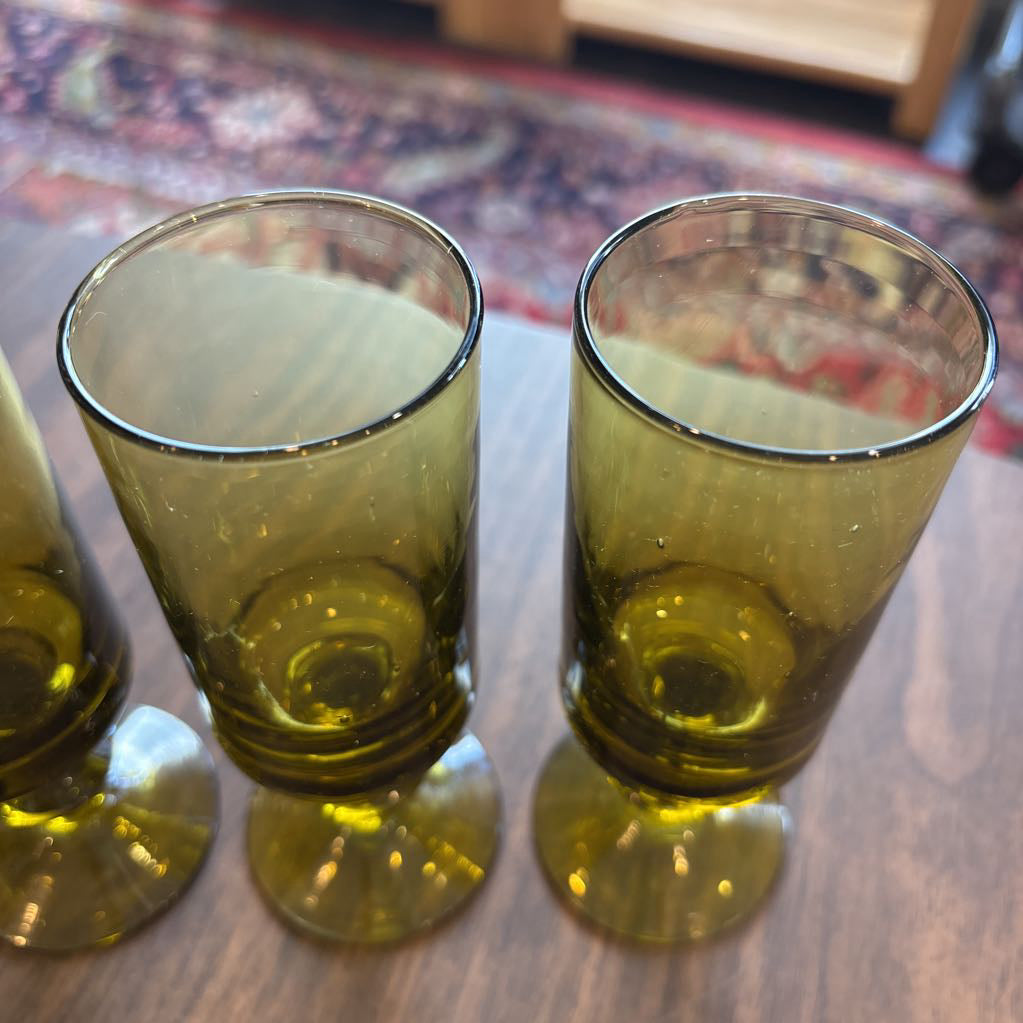 A pair of vintage olive green glass tumblers with footed bases, displayed on a wooden tabletop with a blurred patterned rug in the background.