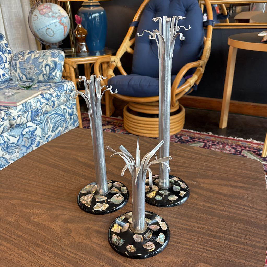 Three silver tree-shaped jewelry stands with circular bases set on a wooden table in a furnished room.