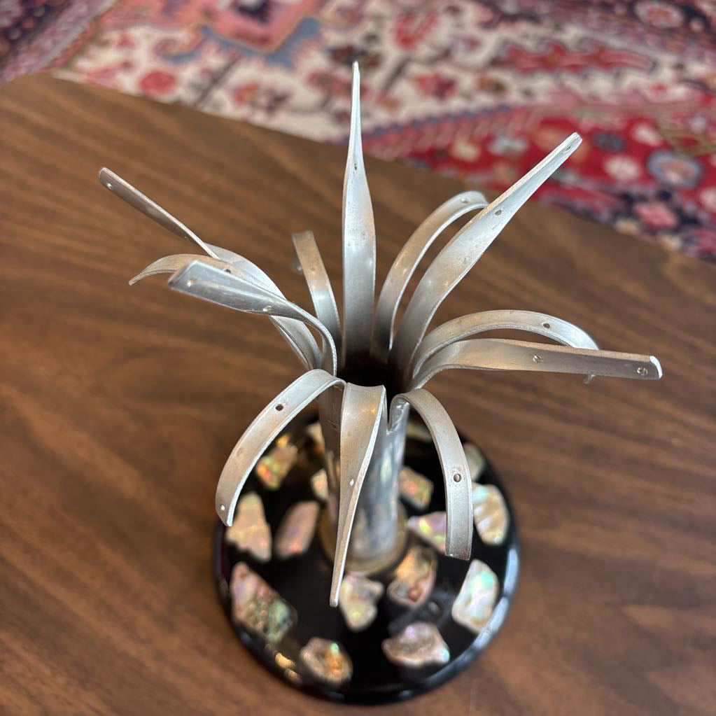 A metallic jewelry tree with curved silver-toned leaves radiating from a black base decorated with iridescent stones, shown on a wooden tabletop.