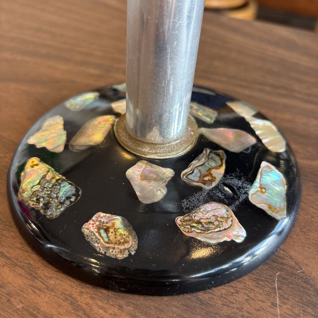 A close-up of a black circular base decorated with irregular iridescent abalone shell pieces, with a silver-toned metal pole rising from the center, set on a wooden surface.