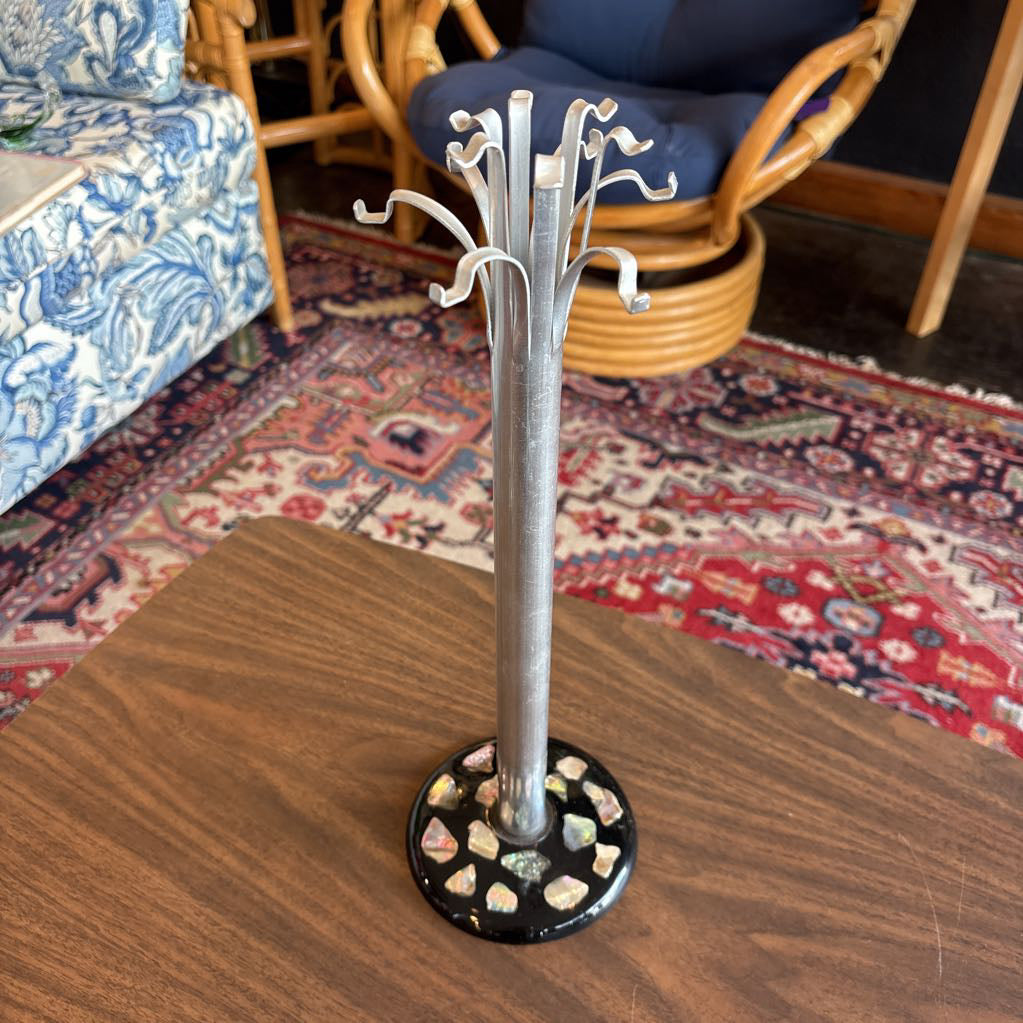 A silver metal jewelry stand with multiple curved hooks on a round black base decorated with small iridescent stones, shown standing on a wooden table in a living room setting.