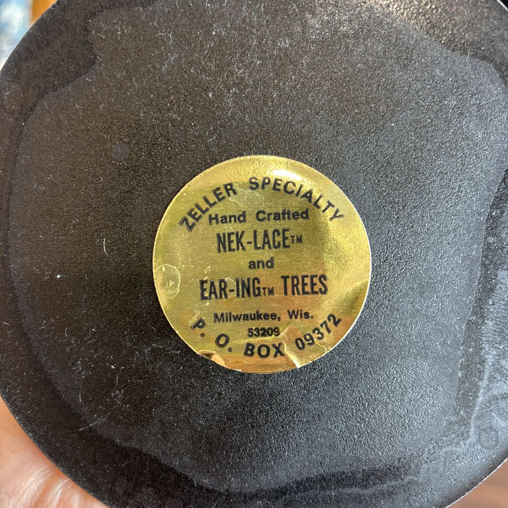 A close-up of a black circular product surface with a round gold label reading 'ZELLER SPECIALTY Hand Crafted NEK-LACE and EAR-ING TREES Milwaukee, Wis. 53206 BOX 09372'.