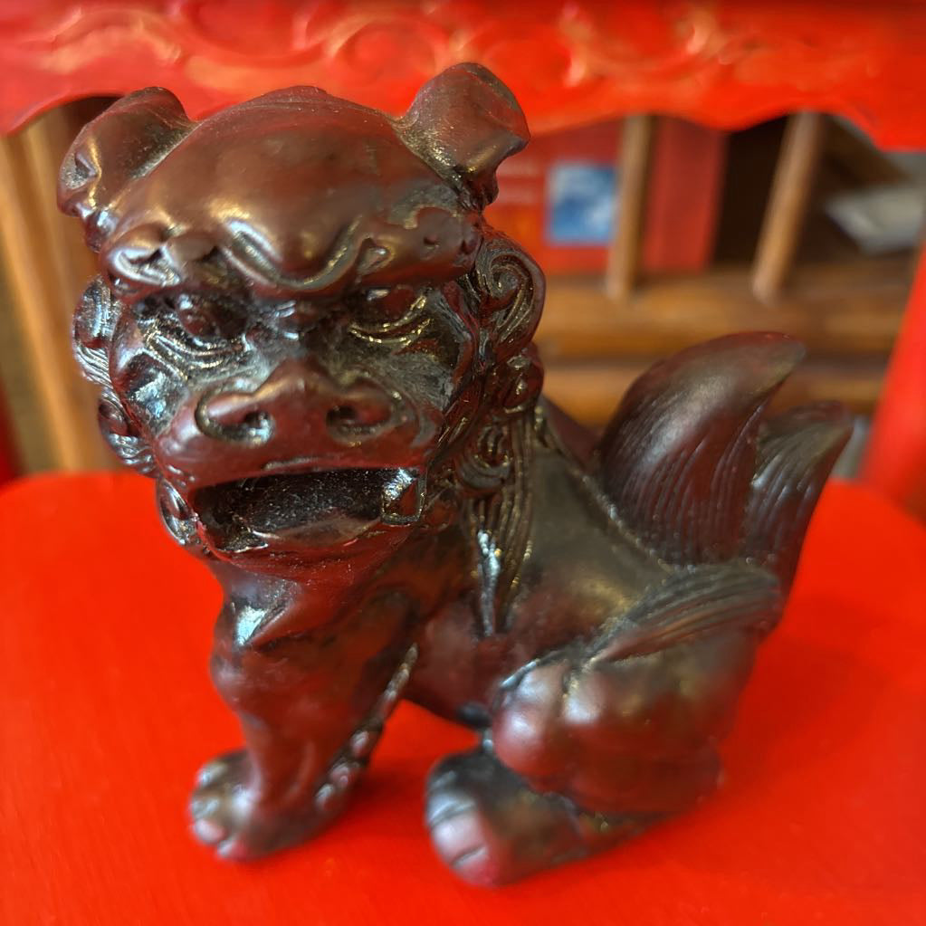 A small dark foo dog statue with an ornate carved face and curled tail, shown on a bright red surface with a blurred red and brown background.