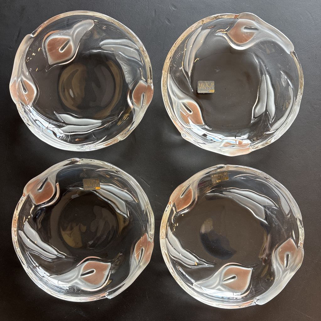 Four glass plates with embossed frosted pink calla lilies and stickers that read 'MIKASA' sitting on a black table.