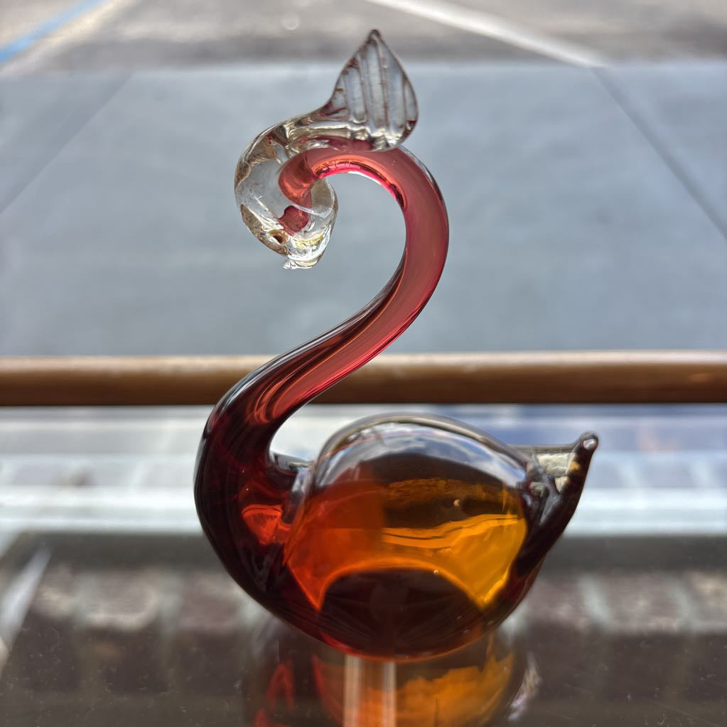 A sculptural glass swan figurine with a curved neck, featuring a deep red glass neck, a clear glass head, and a warm amber-colored glass body, displayed on a reflective surface.