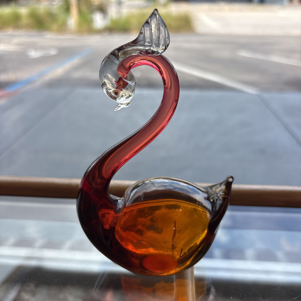 A sculpted glass swan figurine with a translucent amber-orange body and a deep red neck, positioned on a reflective surface with a blurred outdoor background.