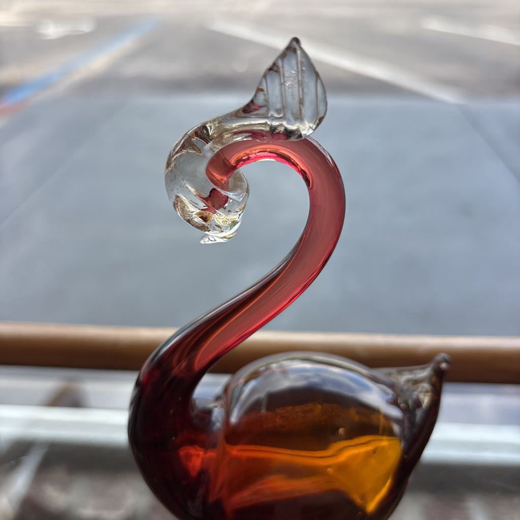 A sculptural glass swan figurine with a curved neck and body, featuring a translucent clear top section, deep red glass along the neck, and amber-colored glass at the base, photographed close-up against a blurred window background.