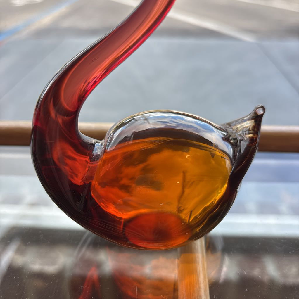 A hand-blown glass decorative object with a curved dark red stem and a bulbous amber-orange glass body, photographed close up against a softly blurred gray background.