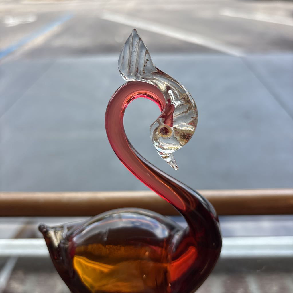 A decorative glass swan figurine with a curved neck, featuring a translucent red body, amber-colored base, and a clear glass head, photographed against a blurred outdoor background.