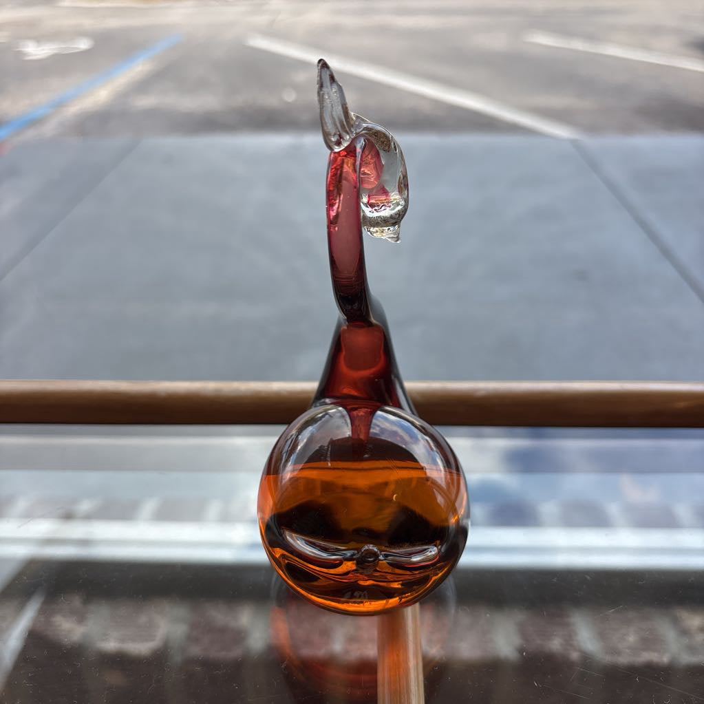 A small hand-blown glass sculpture with a rounded amber-brown base, a curved dark red stem, and a clear glass top, photographed on a reflective surface with an outdoor street scene blurred in the background.