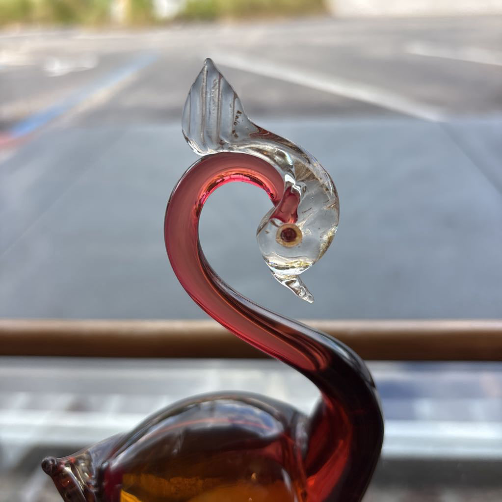 A decorative glass swan figurine with a curved neck and a clear glass fish on its back, photographed in a real environment with a blurred window view in the background.