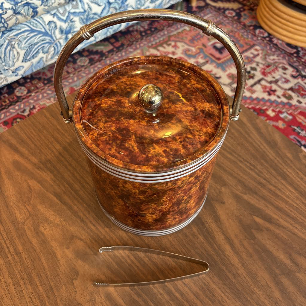A brown speckled ice bucket with white horizontal stripes and the top and bottom and pair of tongs in a room setting.