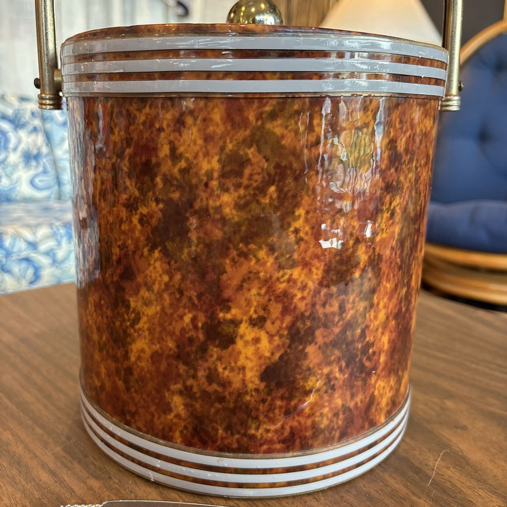 A brown speckled ice bucket with white horizontal stripes and the top and bottom and pair of tongs in a room setting.