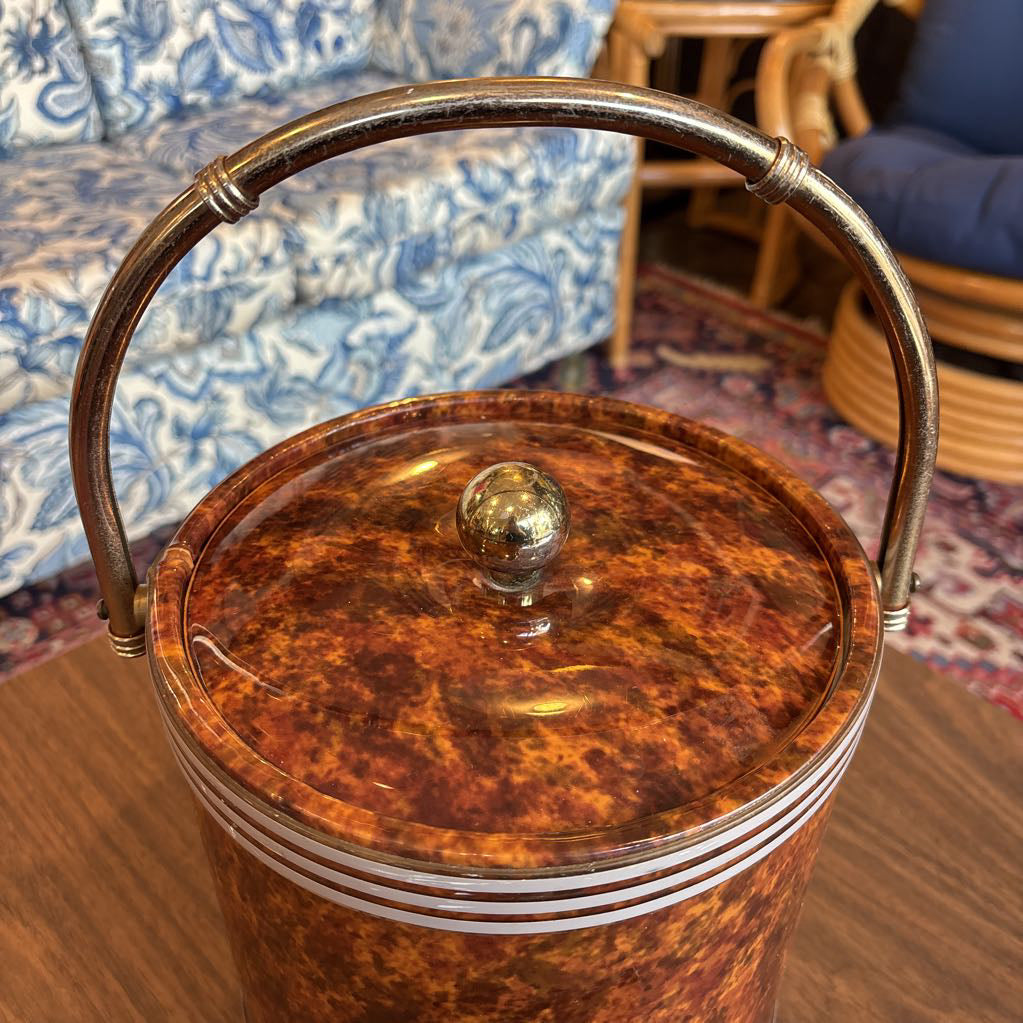 A brown speckled ice bucket with white horizontal stripes and the top and bottom and pair of tongs in a room setting.