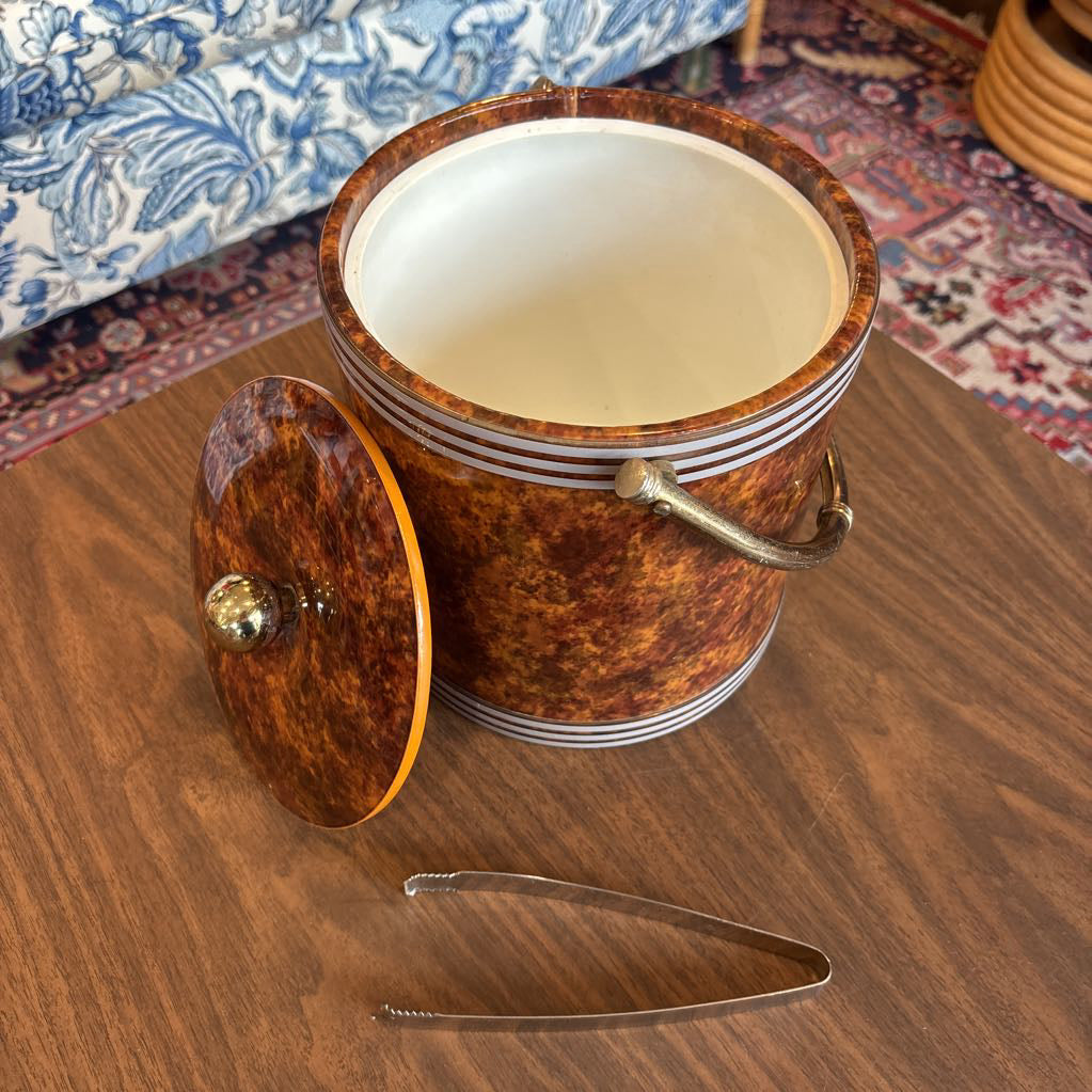 A brown speckled ice bucket with white horizontal stripes and the top and bottom and pair of tongs in a room setting.