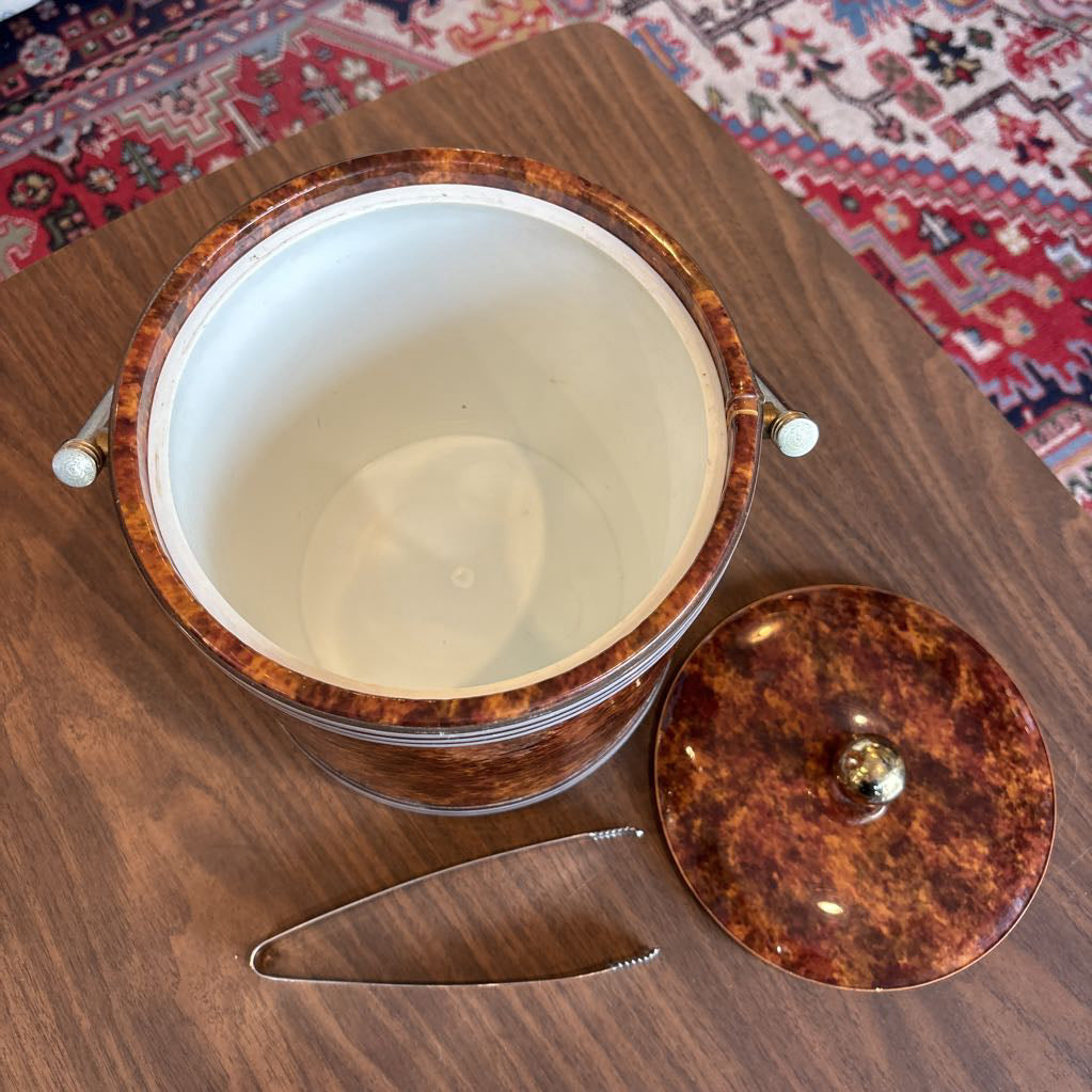 A brown speckled ice bucket with white horizontal stripes and the top and bottom and pair of tongs in a room setting.