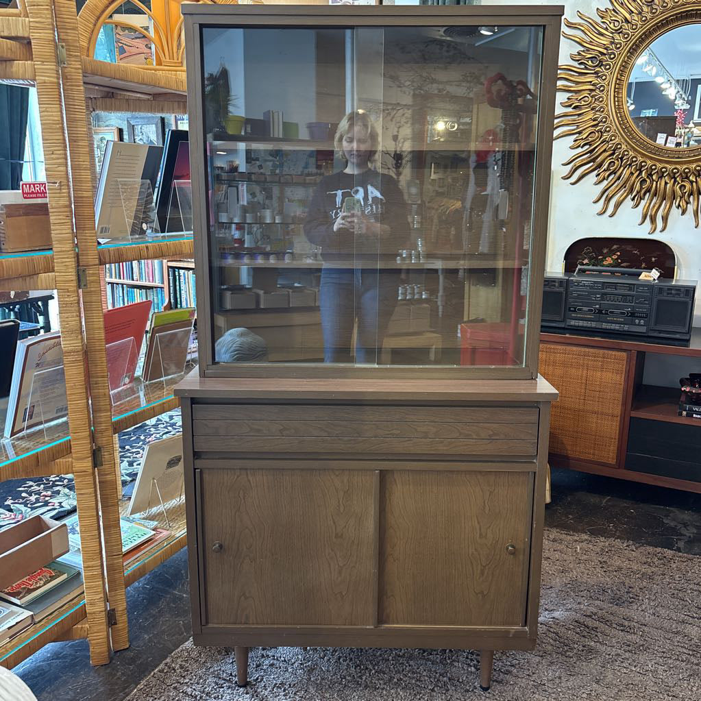 A large wooden hutch with two sliding glass doors and person in the reflection. The cabinet is in a store setting. 