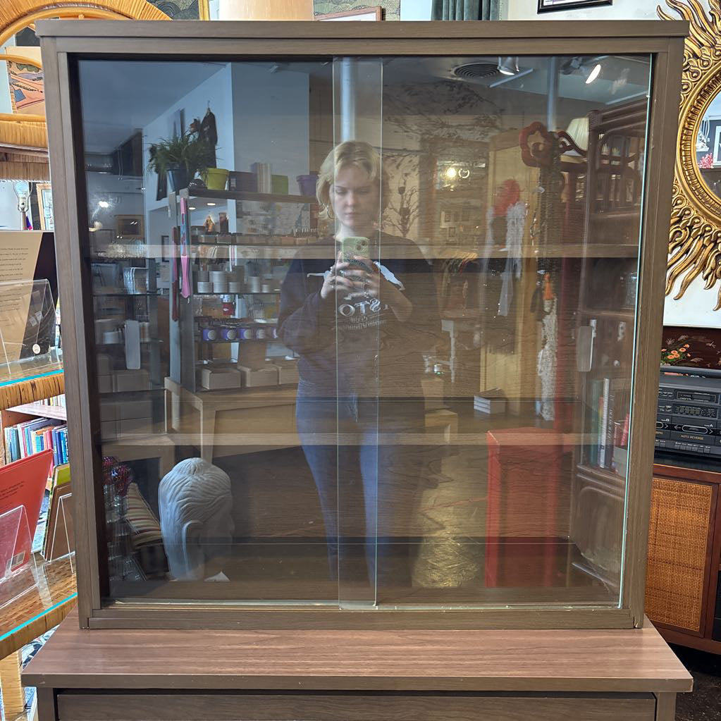 A large wooden hutch with two sliding glass doors and person in the reflection. The cabinet is in a store setting. 