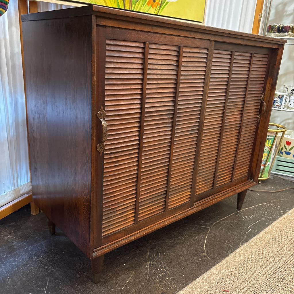 A mid-century modern wooden credenza or sideboard cabinet with louvered doors, shown in a home interior setting.