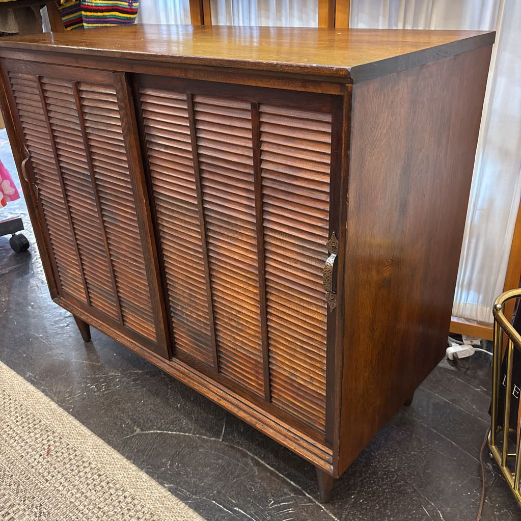 A mid-century modern wooden credenza or sideboard cabinet with two louvered doors and a warm medium-brown wood finish, photographed in an indoor room setting.