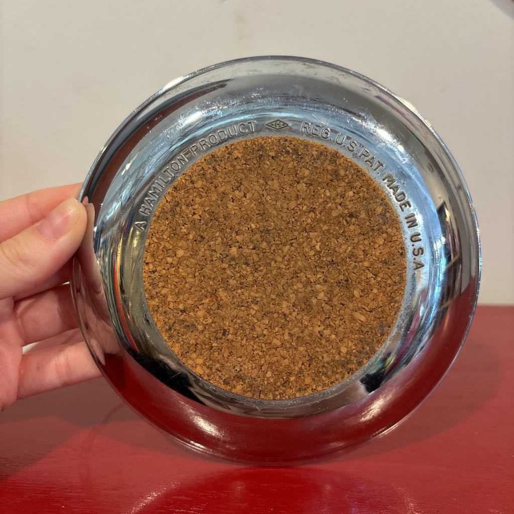 The bottom of a mid-century style chrome ashtray dish on a red surface and up against a white wall. It has branding on the bottom that reads, "A HAMILTON PRODUCT--REG. U.S. PAT. MADE IN U.S.A."