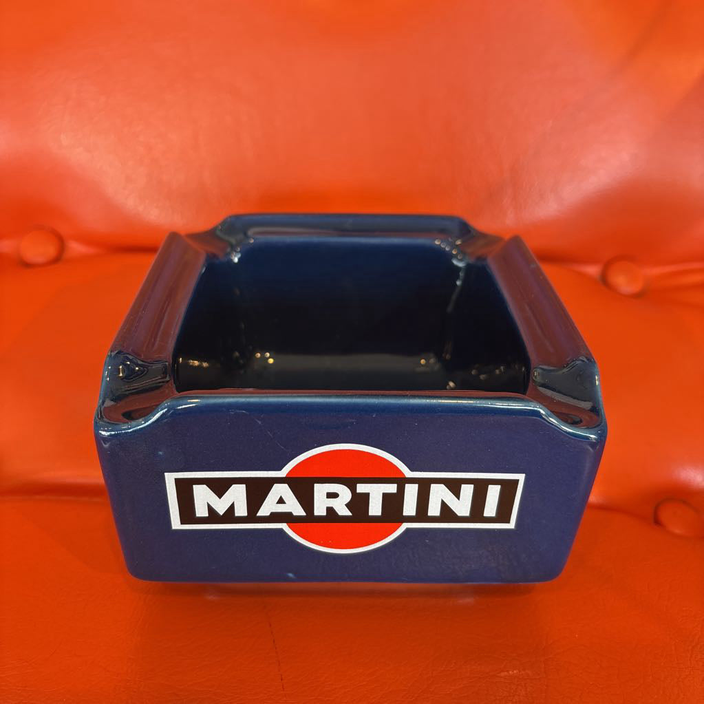 A square blue Martini branded ashtray with a glossy finish, featuring the iconic red and white logo and white 'MARTINI' text on the front, set against a bright orange background.
