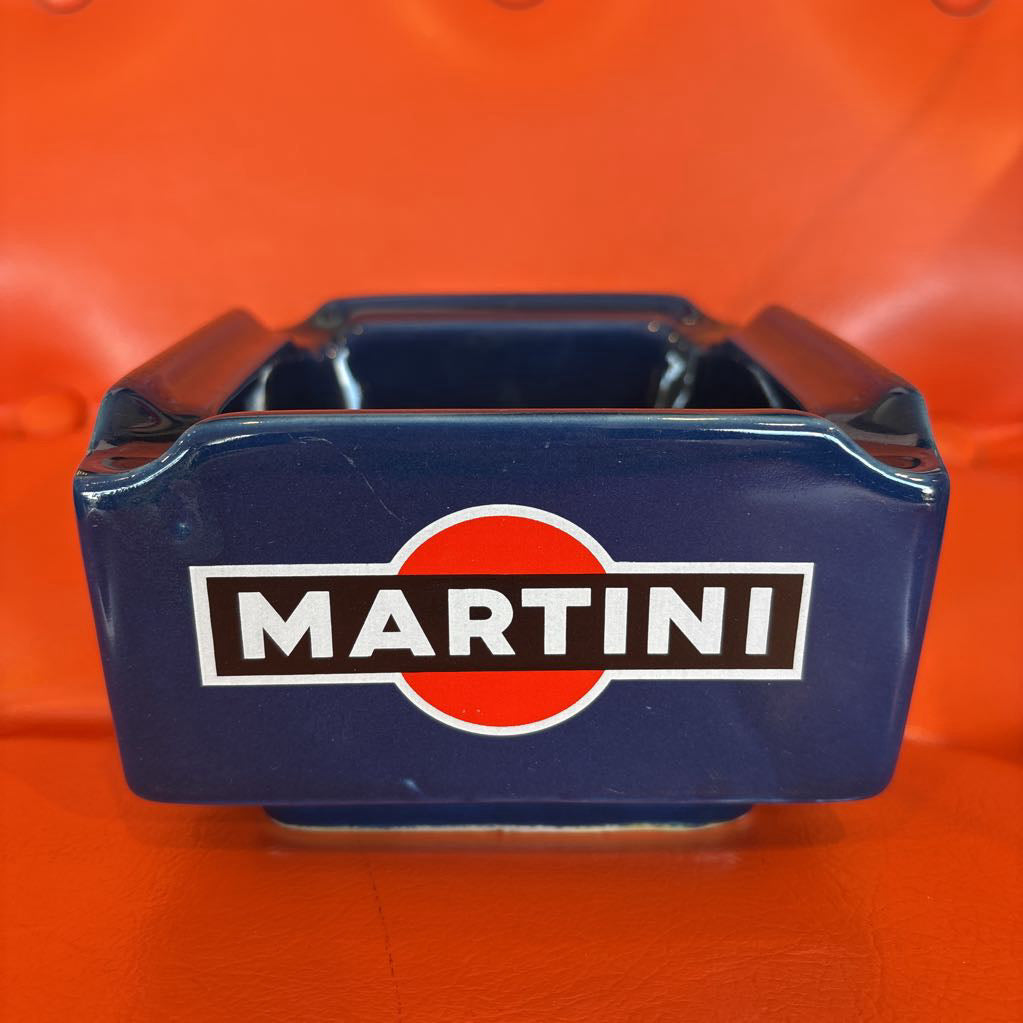 A square blue Martini branded ashtray with a glossy finish, featuring the iconic red and white logo and white 'MARTINI' text on the front, set against a bright orange background.