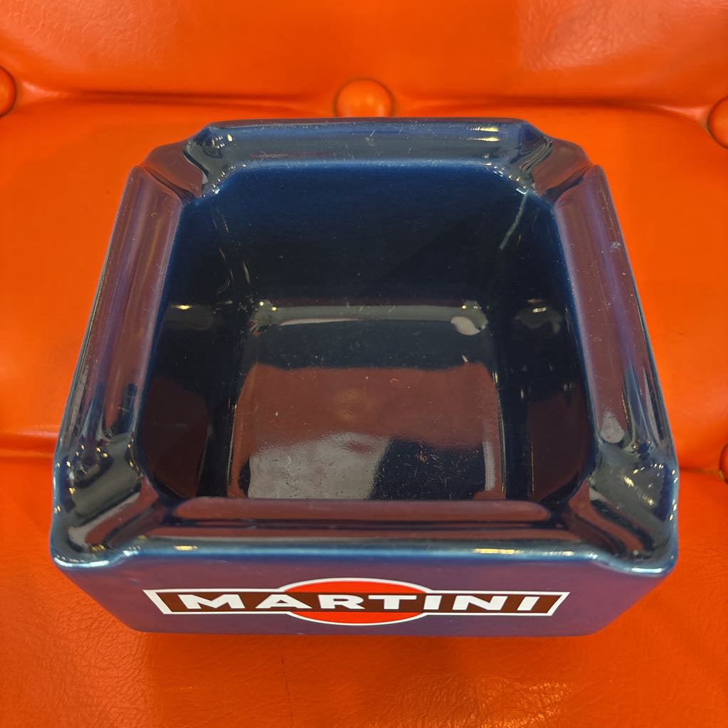 A square blue Martini glass ashtray with a glossy finish and the iconic Martini logo printed on the front, set against a bright orange background.