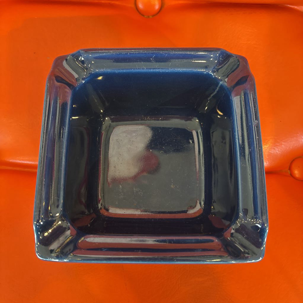 A square glass ashtray with a glossy blue rim and a dark, nearly black interior, shown from a top-down perspective on a bright orange textured background.