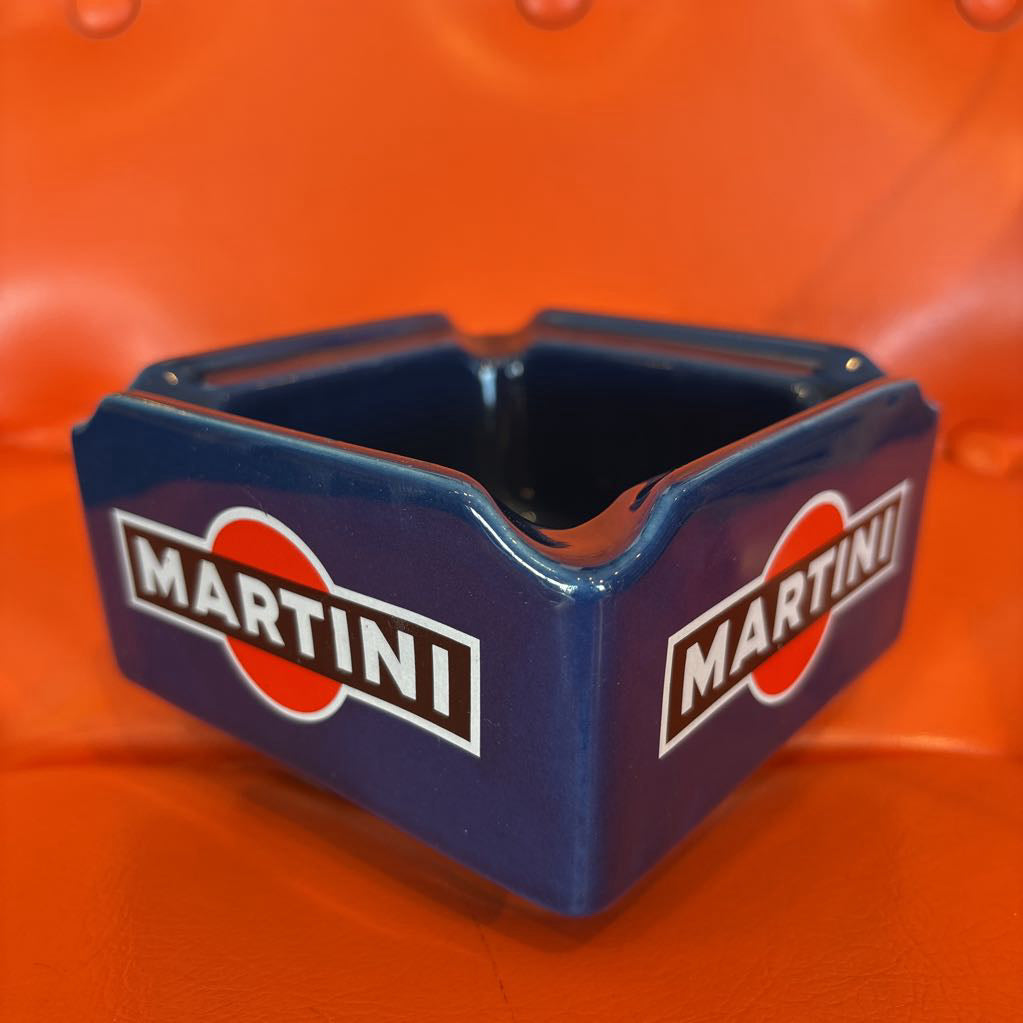 A blue ceramic Martini-branded ashtray with white, red, and black logo graphics, set against a bright orange textured background.