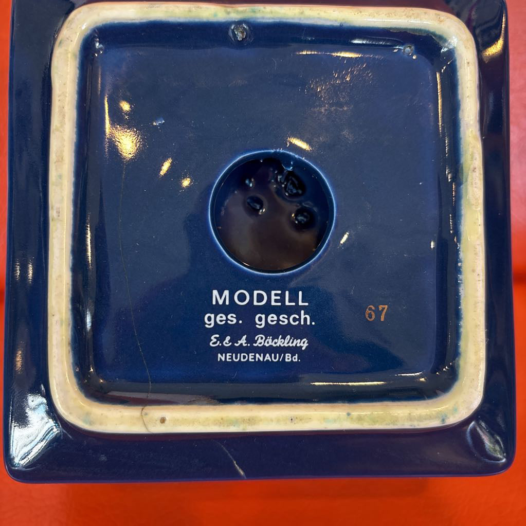 A square dark blue ceramic model dish with a raised cream-colored border and a circular hole in the center, featuring printed text including 'MODELL ges. gesch.