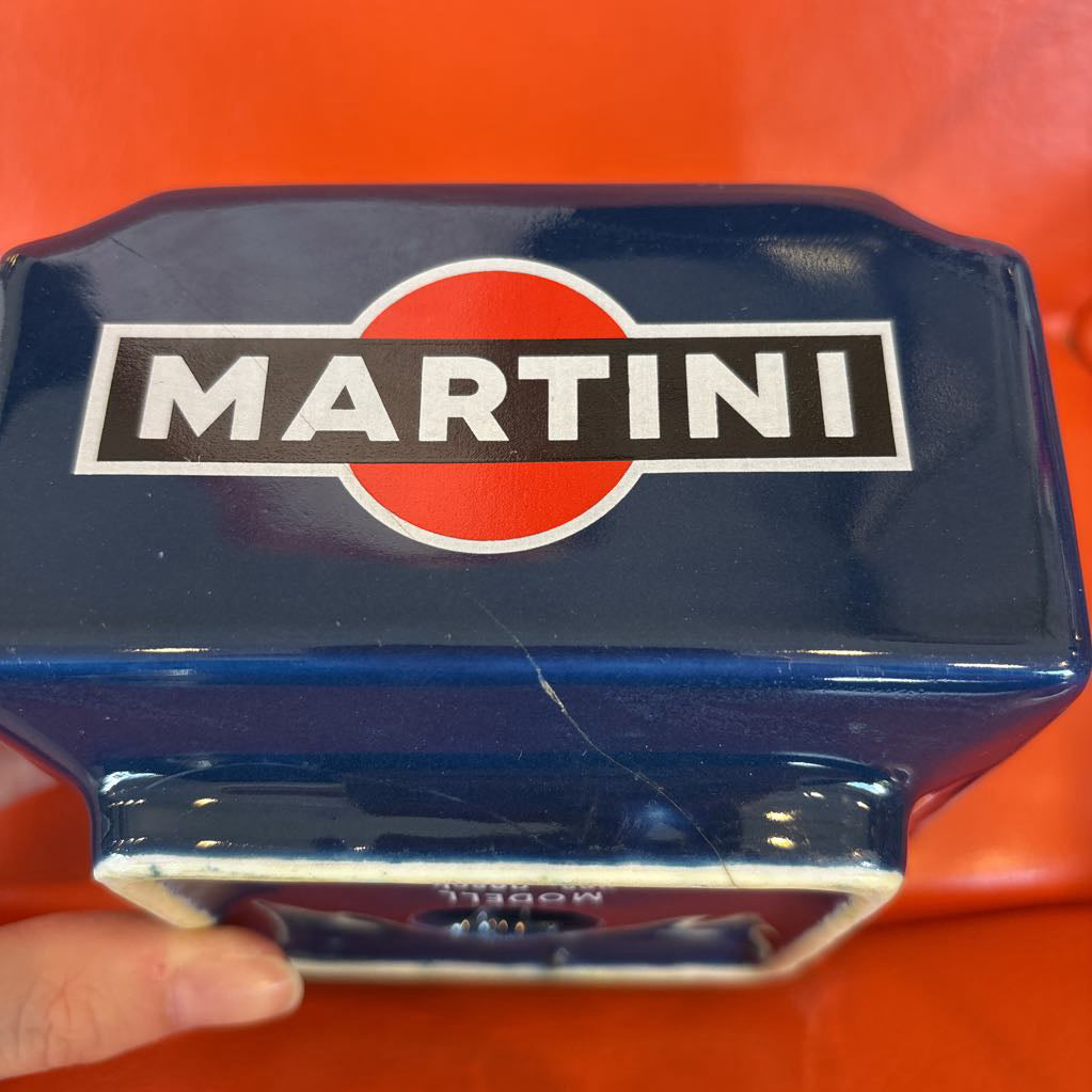 A close-up of a blue Martini glass tumbler with the iconic red and white logo, held by a hand against a bright orange background.