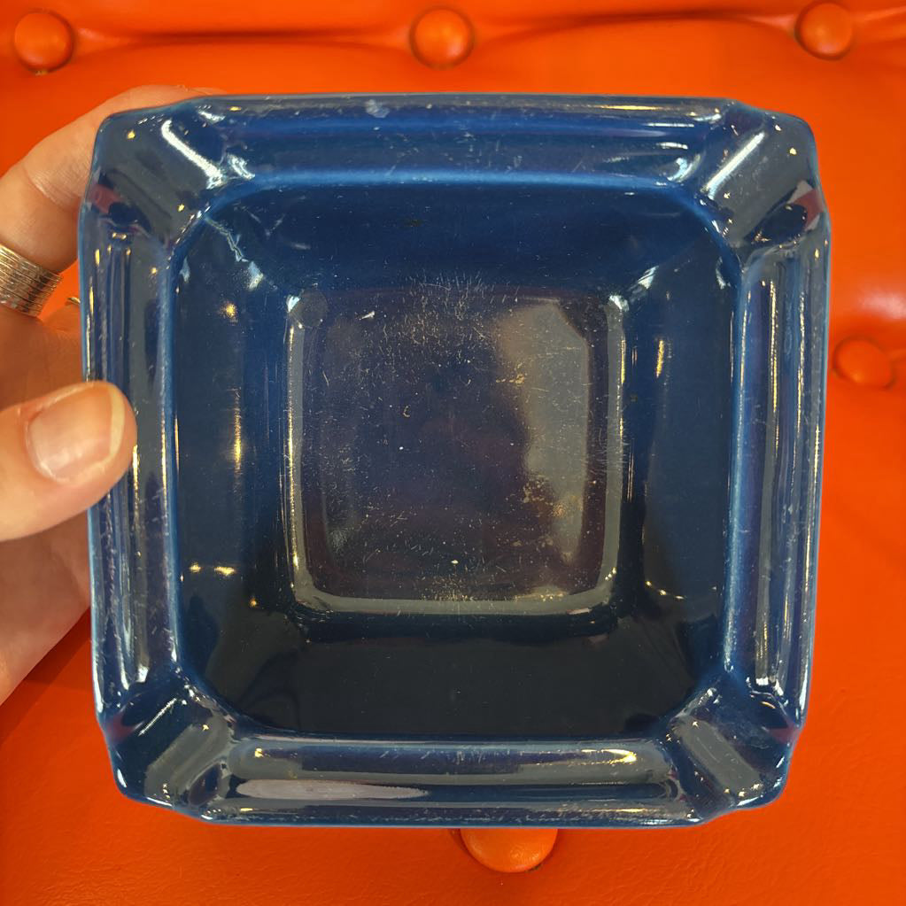 A square blue glass ashtray held by a hand against a bright orange tufted background.