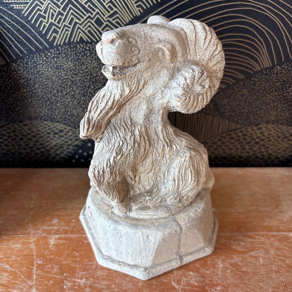 A carved stone figure of a ram on a pedestal. It sits on a wooden surface and a black and gold wallpaper backdrop.